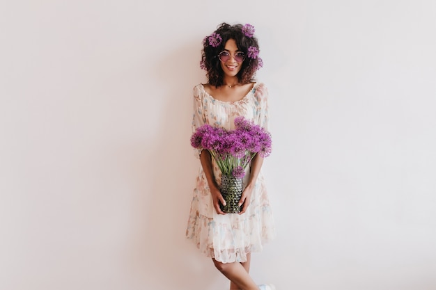 Mulher africana glamorosa com vestido de verão segurando um vaso de flores. Foto interna de inspirada mulher encaracolada, aproveitando a sessão de fotos em casa.