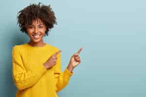 Foto grátis mulher afro alegre aponta para o espaço da cópia, dá passagem ou direção, usa um suéter quente amarelo, tem um sorriso agradável, sente-se otimista, isolada sobre um fundo azul