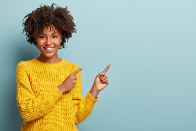 Mulher afro alegre aponta para o espaço da cópia, dá passagem ou direção, usa um suéter quente amarelo, tem um sorriso agradável, sente-se otimista, isolada sobre um fundo azul