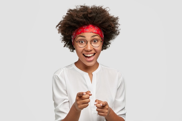 Mulher afro-americana alegre aponta com os dois dedos indicadores, expressa sua escolha, tem cabelo encaracolado e pele escura, usa óculos redondos, camisa casual, isolada sobre a parede branca