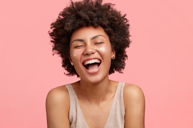 Foto grátis mulher afro-americana alegre com pele escura, ri alegremente, abre muito a boca, tem brilho nas bochechas, fecha os olhos, tem cabelo encaracolado, isolado sobre a parede rosa. conceito de pessoas e felicidade