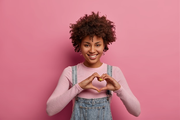 Mulher afro-americana alegre faz um gesto de coração com as mãos, confessa que está apaixonada, sorri positivamente, usa roupa casual, posa contra uma parede rosa pastel. sentimento romântico, conceito de linguagem corporal