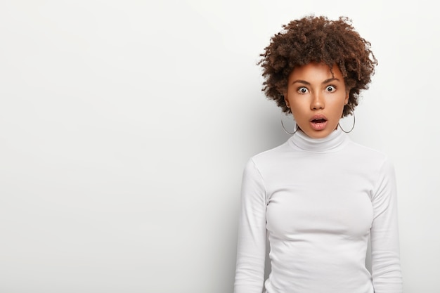 Mulher afro-americana assustada mantém a boca aberta de admiração, tem expressão facial de surpresa assustada, vestida casualmente, isolada na parede branca com espaço em branco no lado direito. conceito de omg.