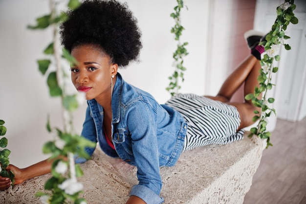 Mulher afro-americana atraente com cabelo afro vestindo saia e jaqueta jeans posada no quarto branco no balanço Modelo preto na moda