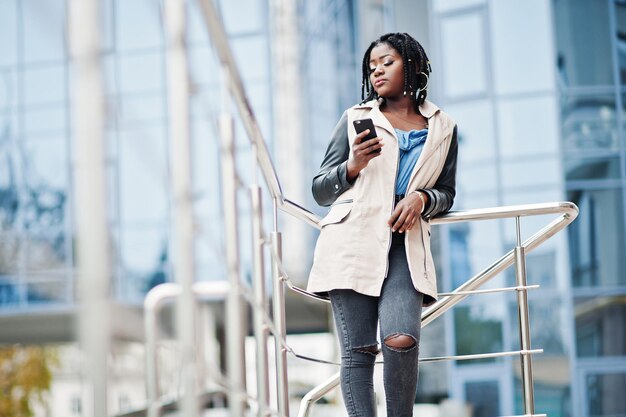Mulher afro-americana atraente com dreads na jaqueta posou perto de grades contra o edifício moderno de vários andares olhando para o celular