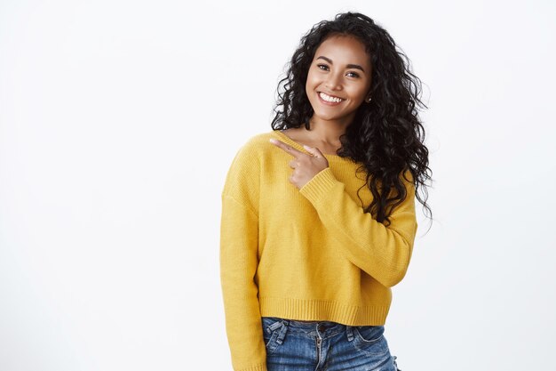 Mulher afro-americana atraente e amigável em um suéter amarelo elegante sorrindo satisfeita, parece confiante e despreocupada, compartilhando notícias legais, apontando para a esquerda, dar conselhos check-out loja legal