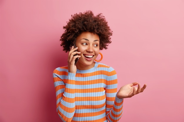 Mulher afro-americana bonita alegre falando ao telefone gosta de conversa desvia o olhar com sorriso fofo mantém a mão levantada usa um macacão listrado casual isolado sobre a parede rosa tem uma conversa interessante