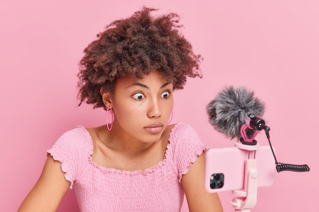 Mulher afro-americana chocada com cabelo encaracolado olha fixamente para a webcam do telefone grava a transmissão ao vivo de vídeo tem cursos de treinamento online