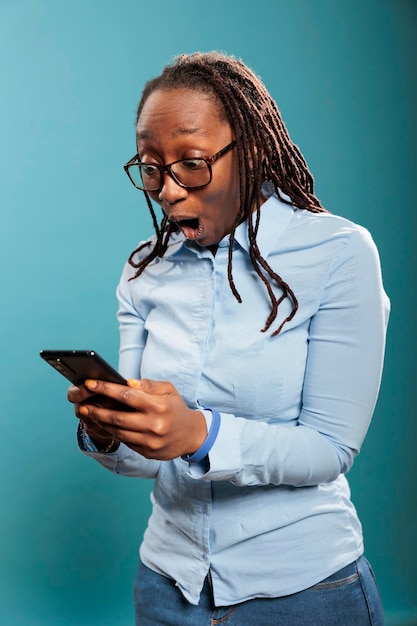 Foto grátis mulher afro-americana chocada com dispositivo de smartphone touchscreen surpreendida pelas mensagens recebidas. surpreso animado jovem adulto com telefone moderno em choque depois de ler as notícias. foto de estúdio