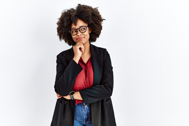 Mulher afro-americana com cabelo afro vestindo jaqueta de negócios e óculos com a mão no queixo pensando em questão, expressão pensativa. rosto sorridente e pensativo. conceito de dúvida.
