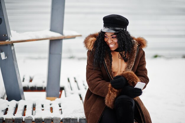 Mulher afro-americana com casaco de pele de carneiro e boné posou no dia de inverno contra um fundo nevado sentado no banco com o telefone à mão