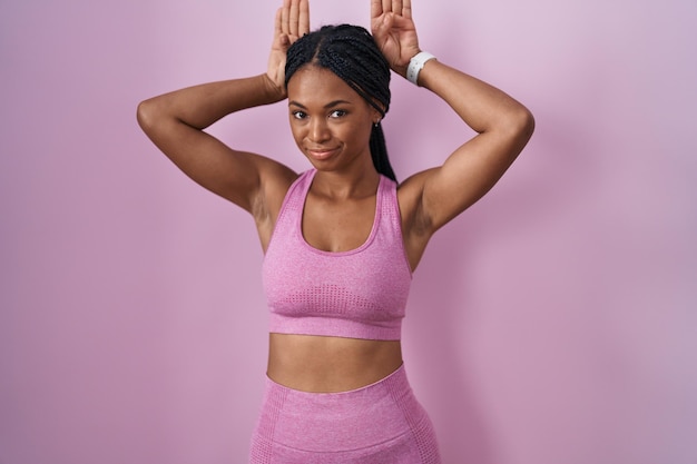 Mulher afro-americana com tranças vestindo roupas esportivas sobre fundo rosa fazendo gesto de orelhas de coelho com as palmas das mãos parecendo céticas e céticas. conceito de coelho de Páscoa.