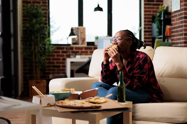 Mulher afro-americana comendo hambúrguer de entrega de fast food, assistindo filme no programa do canal de tv. Servindo refeição para viagem para desfrutar de atividade de lazer com filme na televisão.