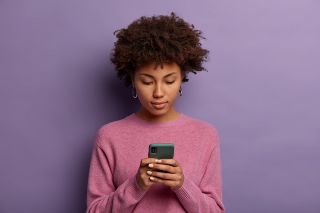 Mulher afro-americana de aparência séria lê mensagem em um celular moderno, navega nas redes sociais, concentra o olhar na tela e usa um macacão casual