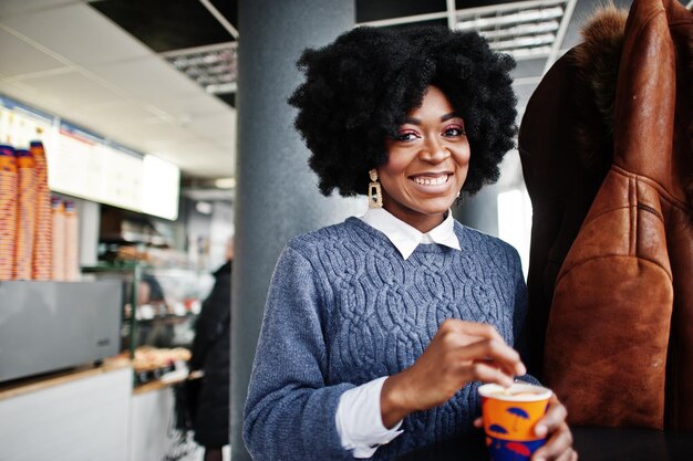 Mulher afro-americana de cabelo encaracolado usa suéter posado no café interior com xícara de chá ou café