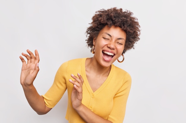 Mulher afro-americana de cabelos cacheados feliz e feliz dançando e sorrindo despreocupada, mantendo os olhos fechados, vestida com um macacão amarelo casual isolado na parede branca