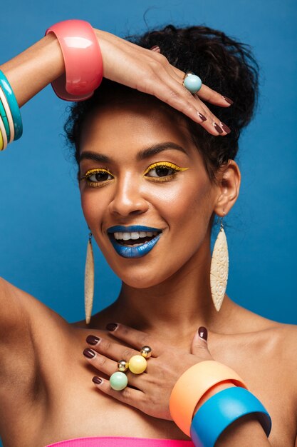 Mulher afro-americana de moda vertical com cosméticos coloridos, mostrando as mãos com acessórios na câmera isolada, sobre parede azul