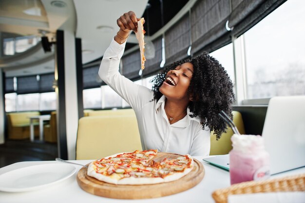 Mulher afro-americana de negócios alegre com cabelo afro usa blusa branca sentada à mesa trabalhando com laptop no café come pizza e bebe coquetel de milk-shake rosa
