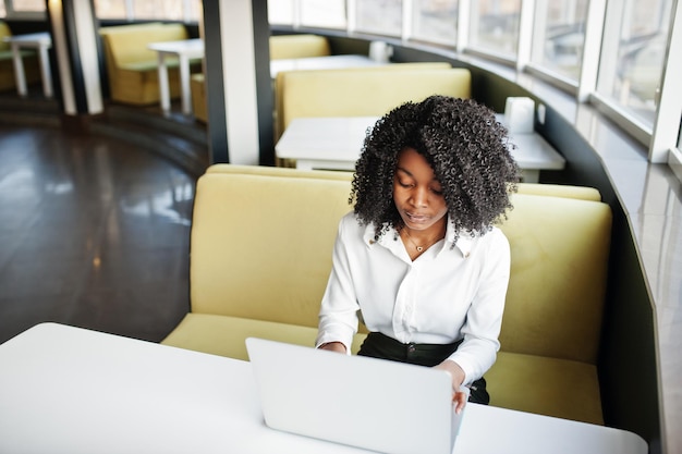 Mulher afro-americana de negócios transparente com cabelo afro usa blusa branca sentada à mesa e trabalha com laptop no café