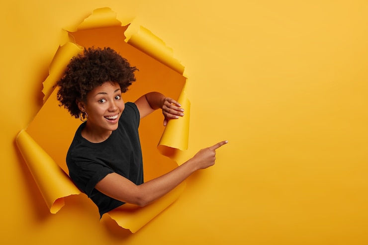 Foto grátis mulher afro-americana de pele escura satisfeita em pé em um espaço rasgado, rindo alegremente e posando em um buraco de papel