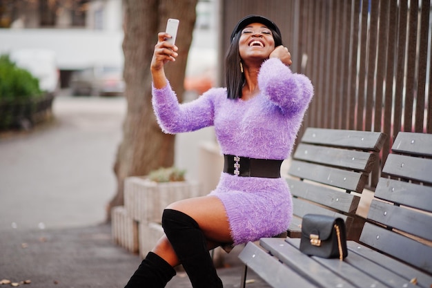 Mulher afro-americana de vestido violeta e boné posou ao ar livre sentado no banco e fazendo selfie no telefone