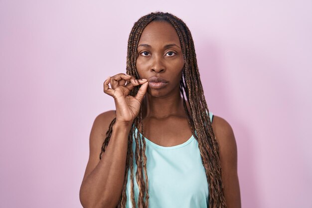 Mulher afro-americana em pé sobre a boca de fundo rosa e lábios fechados como zíper com dedos secretos e tabu silencioso falando