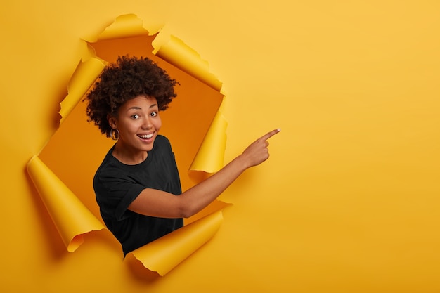 Mulher afro-americana em uma camiseta preta, parada no buraco do papel, apontando para o espaço em branco, parada no papel rasgado