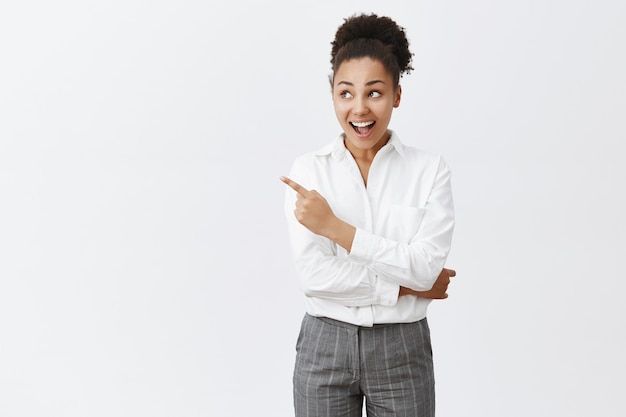 Foto grátis mulher afro-americana empolgada de camisa branca, olhando e apontando para o canto superior esquerdo espantada