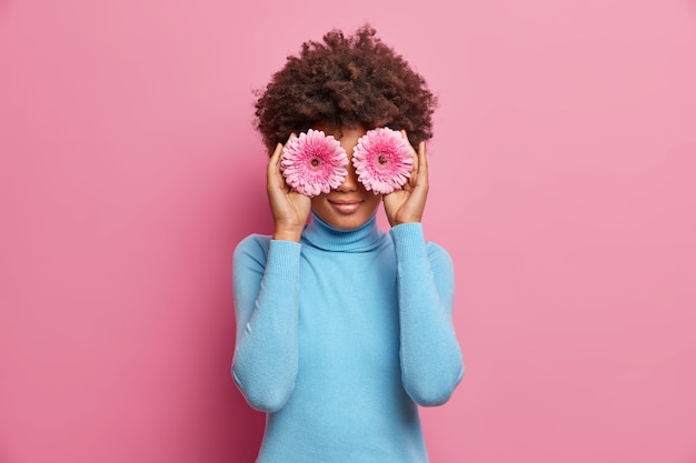 Foto grátis mulher afro-americana encantadora de beleza natural, segurando duas gérberas nos olhos, vestida com gola olímpica azul, poses