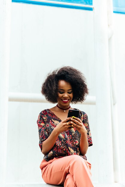 Foto grátis mulher afro-americana encantadora feliz, olhando para a tela do smartphone, sorrindo alegremente