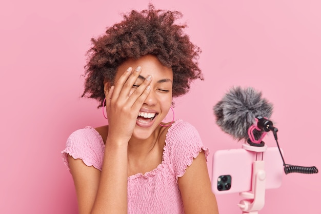 Mulher afro-americana encaracolada e alegre rindo e alegremente mantendo a mão no rosto tem uma conversa engraçada com os assinantes posam contra um fundo rosa