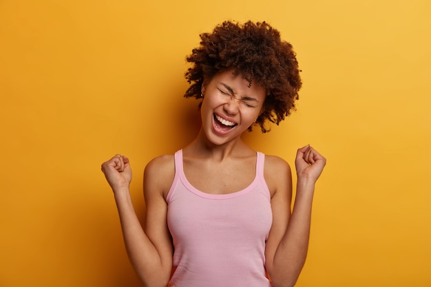 Foto grátis mulher afro-americana encaracolada e animada fecha os punhos em triunfo, comemora boas notícias, inclina a cabeça e exclama de alegria, sendo a sortuda vencedora, vestida com roupa casual, isolada sobre a parede amarela