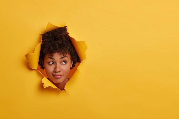 Mulher afro-americana encaracolada olha com expressão curiosa à parte, nota algo interessante, tem beleza natural, isolada sobre fundo amarelo