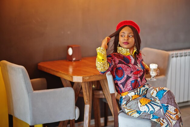 Mulher afro-americana entusiasmada com roupa colorida da moda com boina vermelha relaxando no café aconchegante com uma xícara de café com leite quente nas mãos