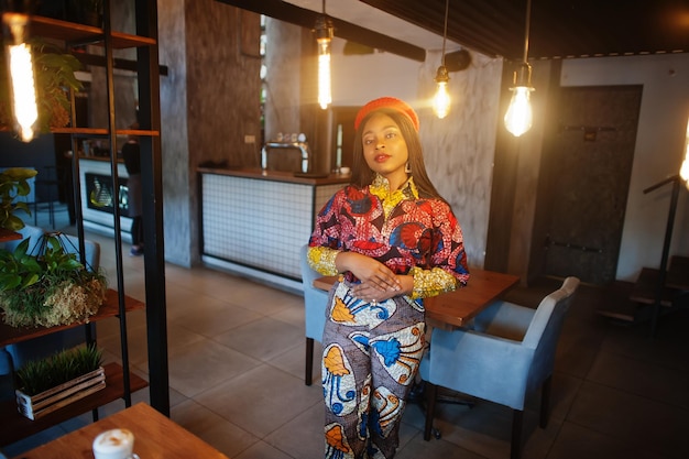 Mulher afro-americana entusiasmada em roupas coloridas da moda com boina vermelha relaxando em um café aconchegante com muitas lâmpadas