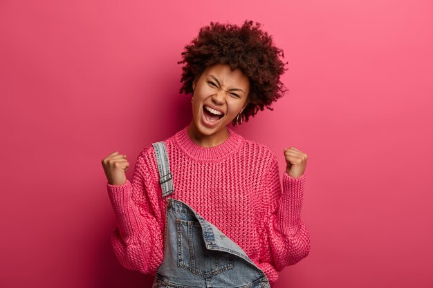 Mulher afro-americana exultante e jubilosa celebra a vitória, alcança o sucesso, exclama com triunfo, veste um suéter, inclina a cabeça, posa sobre uma parede rosa. sendo feliz vencedor. Sim, consegui!