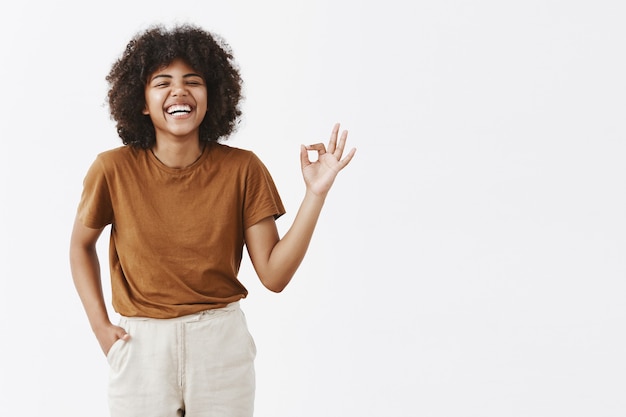 Foto grátis mulher afro-americana feliz e despreocupada com penteado encaracolado desfrutando de uma ótima companhia, rindo alto e se divertindo, mostrando um sinal de ok ou perfeito com os dedos circulados