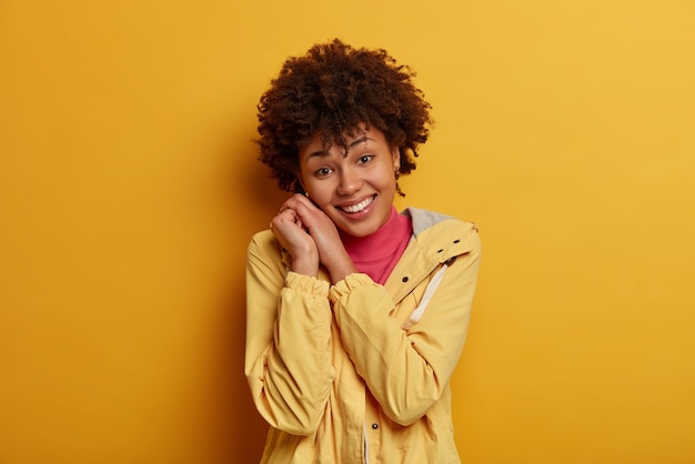 Mulher afro-americana feliz e positiva sorri gentilmente, inclina a cabeça sobre as mãos, feliz em ouvir elogios, vestida de anoraque, observa algo agradável, fica alegre expressa entusiasmo e positividade