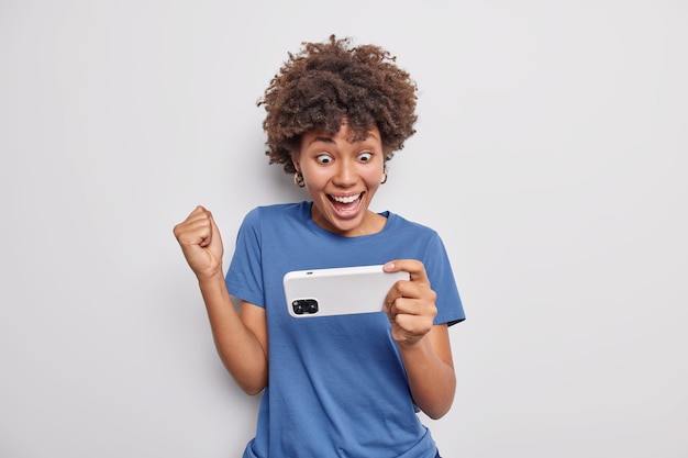 Foto grátis mulher afro-americana feliz vestida com uma camiseta azul casual segura o smartphone horizontalmente, fecha o punho, ganha on-line, assiste ao vídeo isolado sobre a parede branca