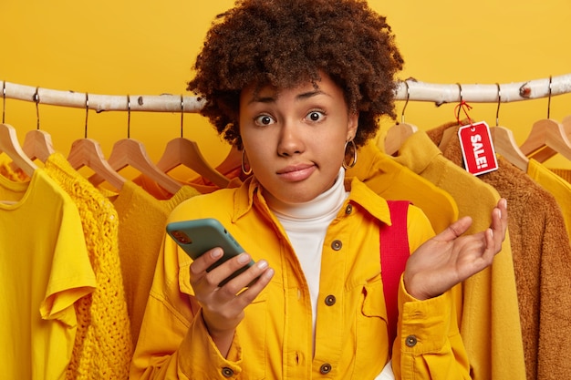 Foto grátis mulher afro-americana hesitante com cabelo encaracolado, levanta a palma da mão, escolhe roupas da nova coleção em butique ou loja de roupas, fica contra vestuários em trapos