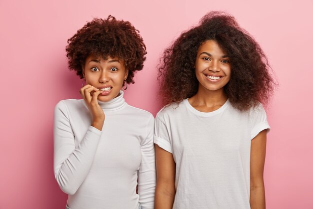 Mulher afro-americana intrigada e nervosa morde as unhas, parece preocupada com a câmera, sua alegre amiga com cabelos crespos e crespos está perto, usa roupas brancas, expressa emoções diferentes