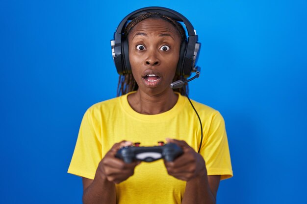 Mulher afro-americana jogando videogame com medo e chocada com surpresa e expressão espantada, medo e rosto animado.