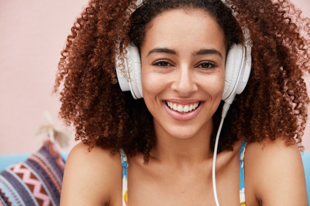 Mulher afro-americana muito feliz com cabelo crespo e espesso aprecia a composição favorita em grandes fones de ouvido modernos