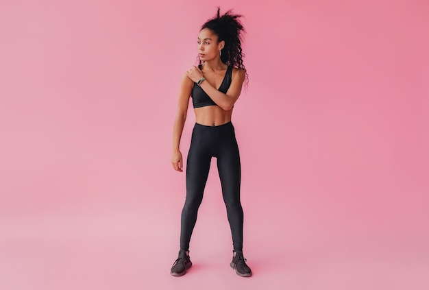 Mulher afro-americana negra atraente em leggings pretas e roupa de fitness superior em fundo isolado rosa confiante cabelo encaracolado estilo esporte corpo atlético