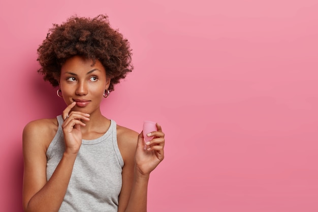 Mulher afro-americana pensativa mantém produto menstrual seguro eficaz e acessível, boa proteção contra vazamentos, escolhe copo menstrual reutilizável, pensa em como usá-lo, espaço vazio em branco na parede rosa