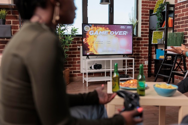 Mulher afro-americana perdendo a competição de videogames, triste e frustrada com o jogo de estratégia de tiro perdido. Pessoa com raiva de perder o jogo, bebendo garrafas de cerveja.