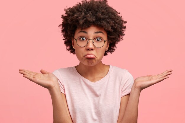 Mulher afro-americana perplexa e hesitante com expressão sem noção, curvas do lábio inferior, ombros encolhidos, vestida com roupa casual, posa contra a parede rosa. Mulher de pele escura não pode tomar decisões