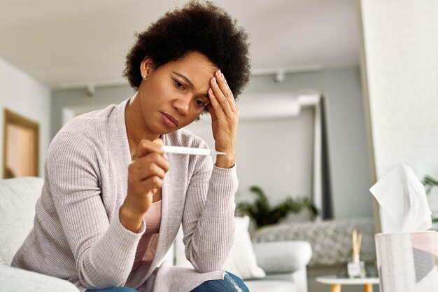 Mulher afro-americana perturbada com dor de cabeça medindo sua temperatura em casa