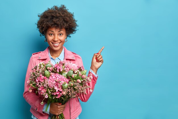Mulher afro-americana positiva com sorriso cheio de dentes indica que ao lado segura um buquê de flores