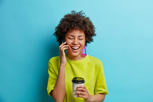 Mulher afro-americana positiva falando pelo celular tem conversa animada segura café para viagem e está de bom humor